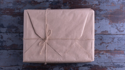 A large classic-looking package wrapped in kraft paper and tied with natural jute twine, on an old wooden surface with a worn gray-blue paint. Packing, gifts, delivery