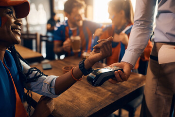 Close up of black woman paying contactless with smart watch during sports championship in bar.