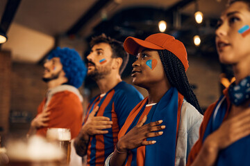 Sports fans holding hand on heart and singing national anthem while watching world championship in bar.