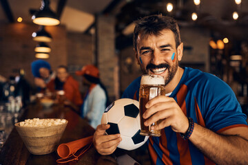 Happy soccer fan drinks beer in bar and looking at camera.