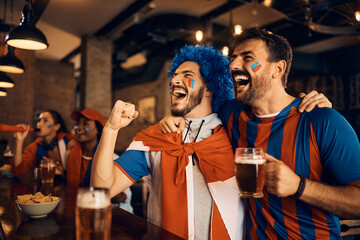 Cheerful soccer fans drinking beer and cheering while watching game on TV in pub.