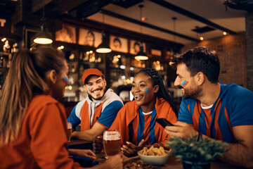 Happy black sports fan and her friends talking while drinking beer in pub during football world cup.