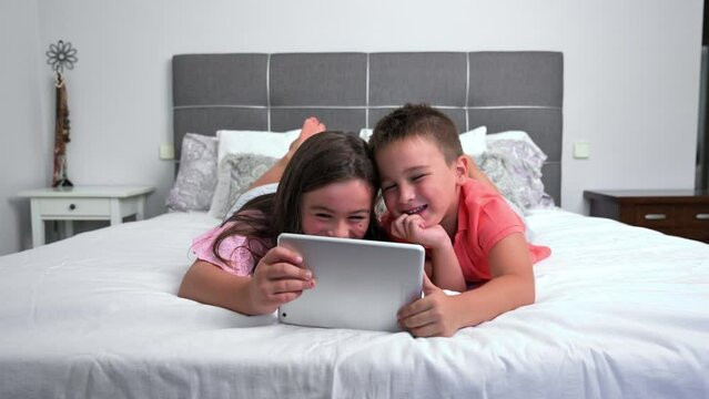 Cute little children with digital tablet using internet at home. Caucasian little sister and brother siblings surfing Internet, watching animated cartoons online on the bed. High quality 4k footage