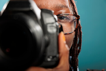 Close up of cheerful smiling professional photographer shooting photos using modern camera while standing on blue background. Young adult woman with photography hobby and DSLR device taking pictures. - Powered by Adobe