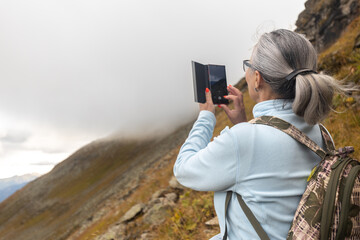 A gray-haired woman in the mountains photographs the fog on a mobile phone. Austria, Alps