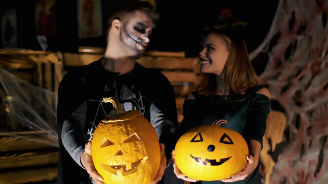date in style of Halloween party, night, twilight, in the rays of light, guy with a girl dressed in costumes and with a terrible makeup are holding two large Halloween pumpkins. High quality photo