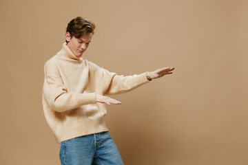 joyful, happy, handsome guy in a long beige sweater poses dancing on a plain background with space...