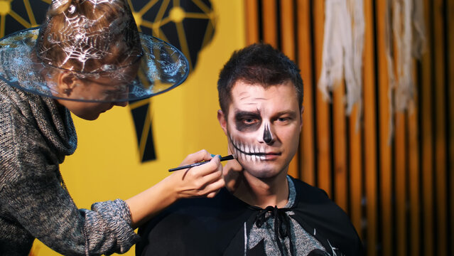 Halloween party, make-up artist draws a terrible makeup on the face of a man for a Halloween party. in the background, the scenery in the style of Halloween is seen. High quality photo