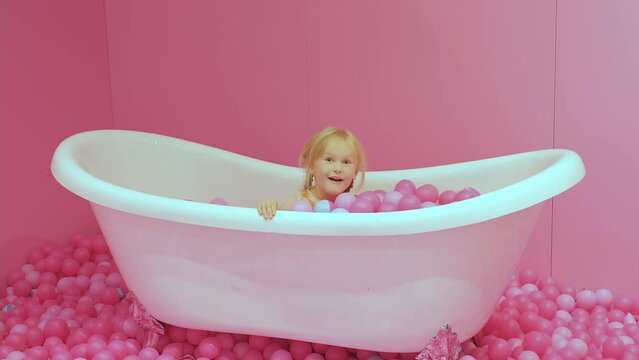 Little pretty blond caucasian girl is lying in the bath with pink plastic balls .