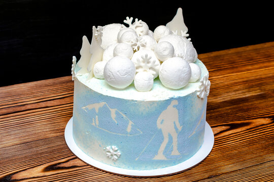 White blue sponge coconut delicious vanilla creamy frosting cake decorated with big round marshmallow, chocolate snowflakes, fir trees,mountain and skier picture,sweet dessert on wooden black table