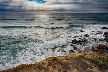 Sun rays shining through the clouds of a  threatening storm as Pacific Ocean waves break over a rocky headland foreshore at Coolum, Sunshine Coast, Queensland.