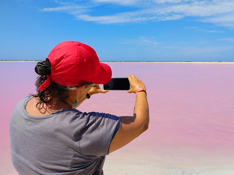 Woman with her back turned takes a picture with her cell phone of the beautiful landscape of pink salt lagoon and blue sky in Las Coloradas, Yucatan, Mexico.
