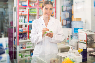 Fototapeta na wymiar Female pharmacist in gown standing behind counter in drugstore. She's writing recipe.