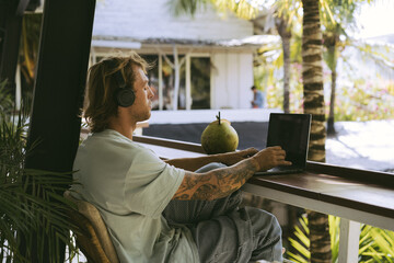 A young smiling man in big headphones works on a laptop in a Bali coworking space. Daily call with...