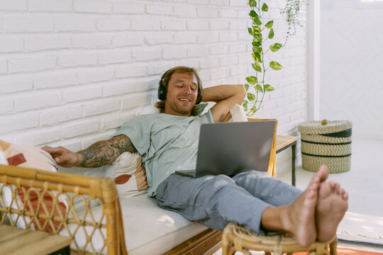 A young man in headphones listens to music, watches a movie on a laptop in a homely cozy atmosphere, sits on the couch.