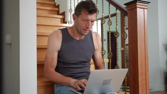 Man mumbling typing on laptop keyboard sitting on stairs in living room. Portrait of adult Caucasian guy messaging online surfing Internet at home indoors. Lifestyle and modern technologies