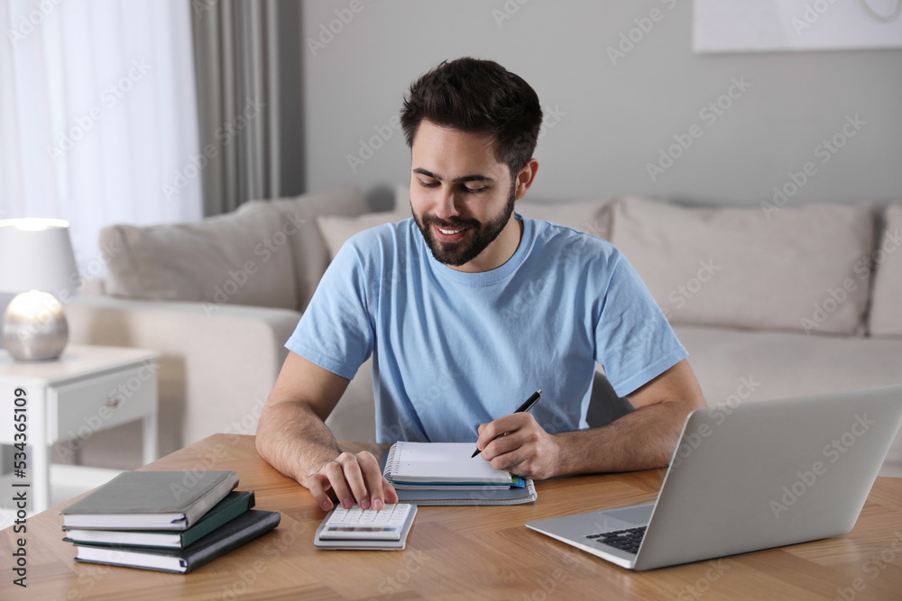 Wall mural Young man counting on calculator during webinar at table in room