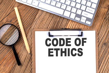 CODE OF ETHICS text on paper clipboard with magnifier and keyboard on wooden background