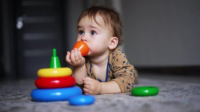 Cute Toddler Boy Lying On The Floor With Toy Pyramid. Lovely Child Took A Toy Top And Put It Into Mouth. Blurred Backdrop.