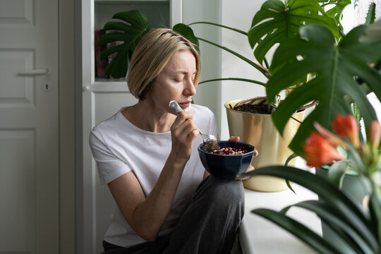 Tranquil Mature Woman Eats Breakfast In Bowl Sitting Alone Near Windowsill With Blooming Houseplant. Unkempt Blonde Middle-aged Female Eats Porridge With Depressive And Sad Expression Closeup