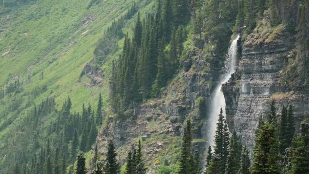 A waterfall in Glacier National park Montana pours down over the cliff past tall pines with green alpine meadows in the distance.