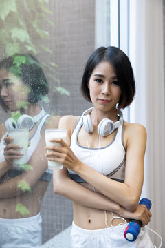 Sporty young woman with glass of milk and dumbbell.  Asian woman in sportswear drinking protein powder milkshake after workout at fitness home gym. Healthcare and weight loss concept.