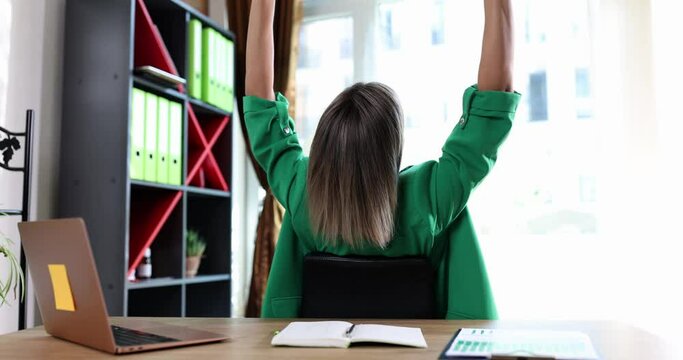 Woman sitting at workplace on chair, bending over, stretching raising hands up, feels happy
