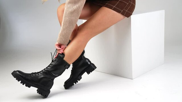 Girl wearing short skirt and high black boots. Model touches the shoe tongue and laces. White backdrop.