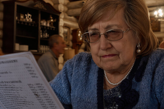 The old lady studies the menu. A grandmother in glasses and a blue jacket sits in a restaurant and carefully reads the menu written on a piece of paper. Selective focus.