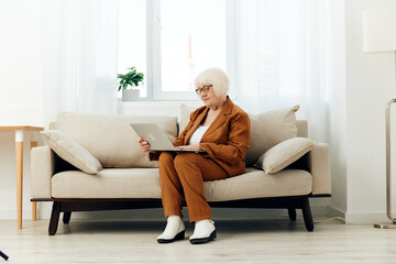 a full-length photo of a nice sweet old lady sitting on a beige sofa in a brown suit working on a laptop