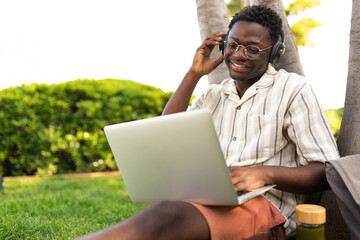 Black man listens to music with headphones and using laptop outdoors. Student doing homework on campus. Copy space.