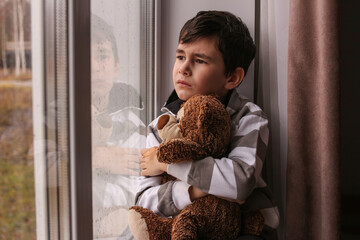 A sad boy looks out the window, hugging a teddy bear. The boy sadly looks out the window at the...