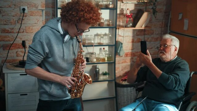 Proud grandfather in a wheelchair takes a video on his phone of  grandson playing the saxophone