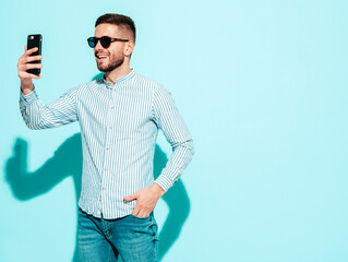 Handsome model. Sexy stylish man dressed in shirt and jeans. Fashion hipster male posing near blue wall in studio. Holding smartphone. Looking at cellphone screen. Using apps. In sunglasses