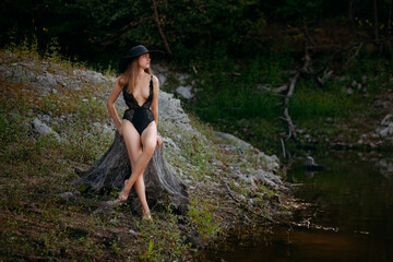 A young girl with long blond hair and a beautiful figure, in black openwork lingerie and a black wide hat, sits on a stump on the banks of a picturesque river.