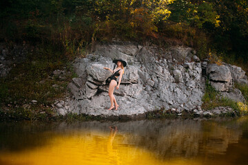 A young girl with long blond hair and a beautiful figure, in black openwork lingerie and a black wide hat, stands against the backdrop of a rock, on the banks of a picturesque river.