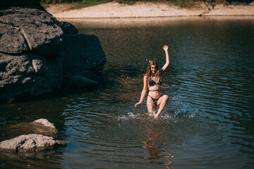 A young girl with long blond hair, a slender figure, in a black swimsuit jumps in the river against the backdrop of rocks and laughs.