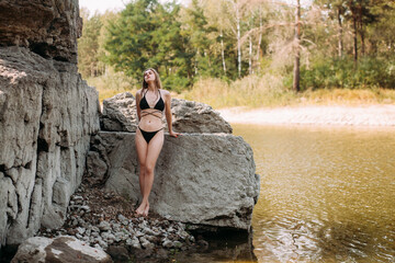A beautiful girl with long blond hair, a slender figure, in a black swimsuit, stands by a rock near the ocean on a hot sunny summer day.
