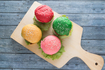 Four colorful mini burgers on red, yellow, green and pink sesame buns served on a wooden board. Top...