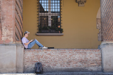 Young man studying with a tablet sitting on a wall at the University of Bologna, Italy. - obrazy, fototapety, plakaty
