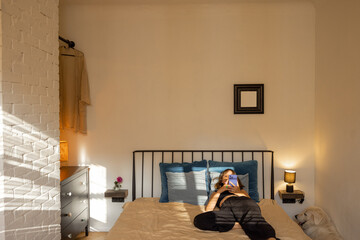 Woman lying with smart phone on bed in beautiful and sunny bedroom. Leisure time and domestic lifestyle