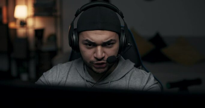 Close-up of bearded guy in cap wearing headphones. Man plays online game, he is finalist and one step away from winning. Guy communicates with opponent. Man is infinitely happy about victory.