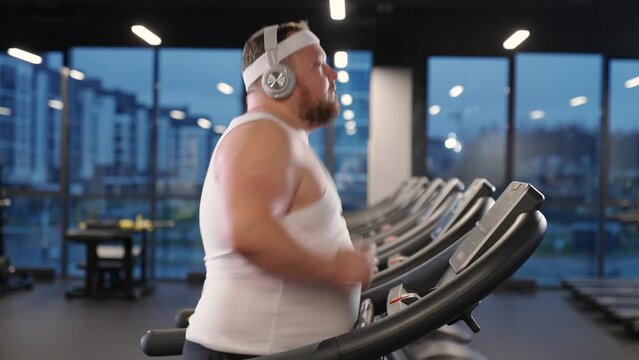 Motivation and self improvement, overweight man run on a treadmill and listening to music with headphones, aerobic exercise and endurance training in the gym, fat burning.
