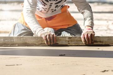 Close up of man builder placing screed rail on the floor covered with sand-cement mix at construction site. Male worker leveling surface with straight edge while screeding floor. Blurred background.