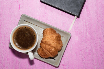 Coffee and croissant in plate on pink table has book top view.