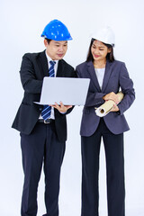 Millennial Asian successful professional male and female engineer in formal suit and safety hard helmet standing together holding blueprint looking at laptop notebook computer on white background