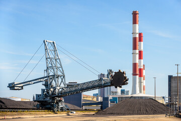 A bucket-wheel stacker-reclaimer and coal stockpiles in a thermal power station with red and white smokestacks on a sunny day.