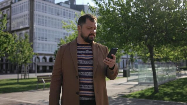 A business man with a beard in his coat looks at the phone ringer in the business district, waiting for a business meeting.