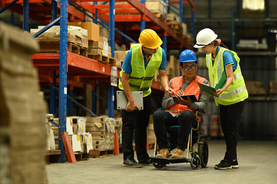 Asian male manager in wheelchair and young workers using digital tablet, inspecting stock product in a large warehouse