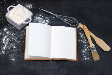 Leeres  Kochbuch auf einem dunklem Schieferhintergrund mit Schneebesen, Kochlöffel und einer...